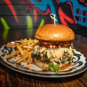 A juicy cheeseburger with lettuce, tomato, onions, and creamy sauce is served on a decorative plate with golden French fries and a small cup of dipping sauce; a colorful graffiti mural is in the blurred background.