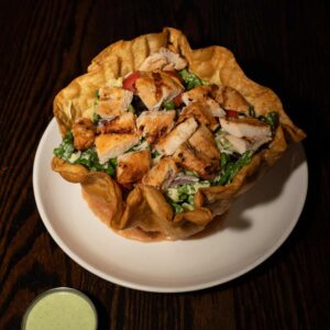A taco salad served in a crispy tortilla bowl filled with chopped grilled chicken, lettuce, tomatoes, black beans, and cheese, on a white plate atop a dark wooden table. A small cup of green dressing sits beside the plate.