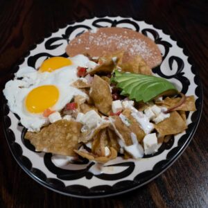 A decorative plate holds sunny-side-up eggs, crispy tortilla chips with salsa, queso fresco, and crema, sliced avocado, and a serving of refried beans dusted with cheese, set on a dark wooden table.