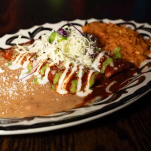 A plate of Mexican food featuring enchiladas topped with crema, green sauce, and shredded cabbage, served with refried beans sprinkled with cheese and a portion of Mexican rice on a decorative black and white plate.