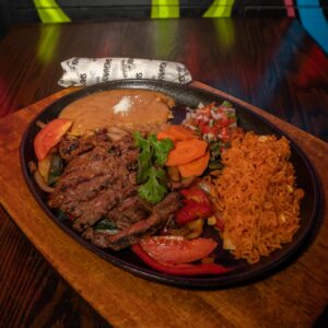 A sizzling platter with grilled steak strips over sautéed onions and peppers, sliced tomatoes, cilantro, Mexican rice, refried beans with cheese, carrots, and fresh pico de gallo. A wrapped flour tortilla sits behind the plate on a dark wooden table.