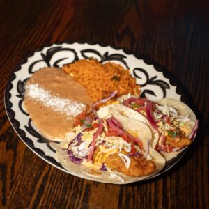A decorative plate holds two fish tacos topped with shredded cabbage, cheese, tomatoes, onions, and cilantro, served with a side of refried beans sprinkled with cheese and a portion of Mexican rice on a dark wooden table.