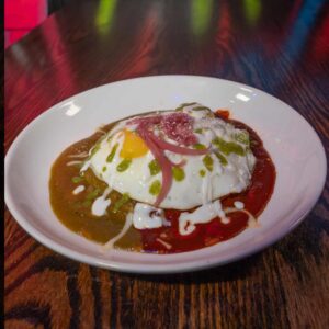 A white plate on a wooden table holds huevos divorciados—two eggs topped with crema, pickled onions, and served with red and green salsas on either side of the eggs, creating a colorful, divided presentation.