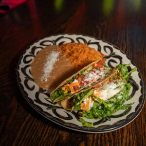 A decorative plate holds two tacos filled with lettuce, tomato, cheese, and sauce, alongside a serving of refried beans topped with cheese and a mound of orange Mexican rice, all set on a dark wooden table.