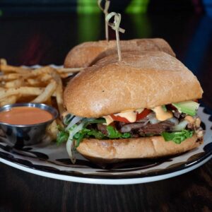 A close-up of a sandwich with lettuce, onions, avocado, tomato, grilled meat, cheese, and sauce on a ciabatta roll, served with a side of golden French fries and a small cup of dipping sauce on a white plate with a black rim.