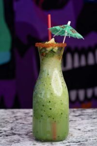 A tall glass bottle filled with green margarita slushie and garnished with diced cucumber, a spicy rim, a red straw, and a small green umbrella on top. The refreshing drink is placed on a marble surface, with a vibrant, colorful background.