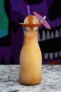 A tall glass bottle filled with a refreshing orange-colored margarita slushie is shown on a marble surface. The bottle is garnished with a lime wedge coated in chili powder and a small pink paper umbrella. The background features a vibrant, abstract mural with bold colors and dark shapes.