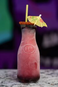 A tall, refreshing pink cocktail is garnished with a small paper umbrella and has a chili-rimmed top. This margarita slushie is filled with a gradient of colors from dark purple at the bottom to light pink at the top and is served on a marble surface against a blurred, colorful background.