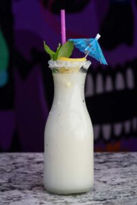 A tall glass bottle filled with a creamy white beverage, topped with mint leaves, a lemon slice, and an umbrella cocktail pick. The rim of the bottle is coated with sugar, making it look like a refreshing margarita slushie. In the background, there’s a colorful, out-of-focus mural.