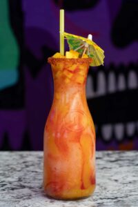 A tall, clear bottle filled with a vibrant mango drink swirls with red chamoy sauce. The refreshing concoction, reminiscent of a margarita slushie, is garnished with diced mango, a small umbrella, and a yellow straw, all resting on a granite countertop with a colorful, abstract background.