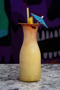 A frosty, creamy beverage served in a clear, tall glass with a chili-rimmed edge, garnished with a slice of pineapple, a blue paper parasol, and a green straw, sits on a white and gray marble surface against a colorful, abstract background.