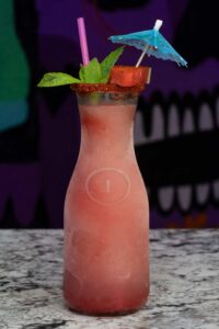 A tall, frosty glass jar filled with a refreshing pink margarita slushie garnished with a chili powder rim, mint leaves, a slice of watermelon, a blue paper cocktail umbrella, and two straws—one pink, one purple. The background shows abstract dark-colored artwork.