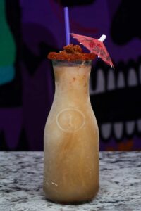 A tall glass bottle filled with a light brown, icy margarita slushie. The rim is coated with reddish seasoning, and a small pink paper umbrella decorates the top. A purple straw pops out from the top, making it look incredibly refreshing against the dark background with abstract shapes.