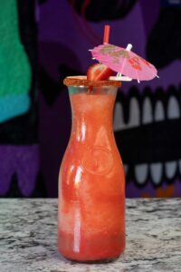 A tall, clear bottle filled with a red, refreshing margarita slushie featuring a salted rim and garnished with a strawberry slice. A pink paper umbrella is also perched on the rim, and the background showcases abstract, colorful art.